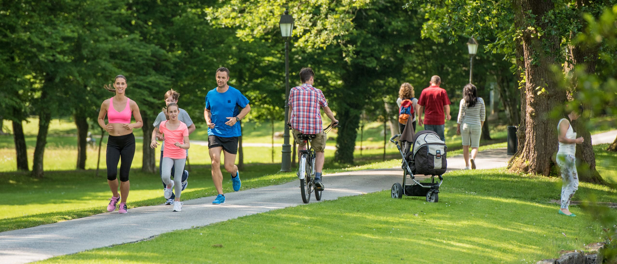 Family jogging and man cycling in park, people walking in background.